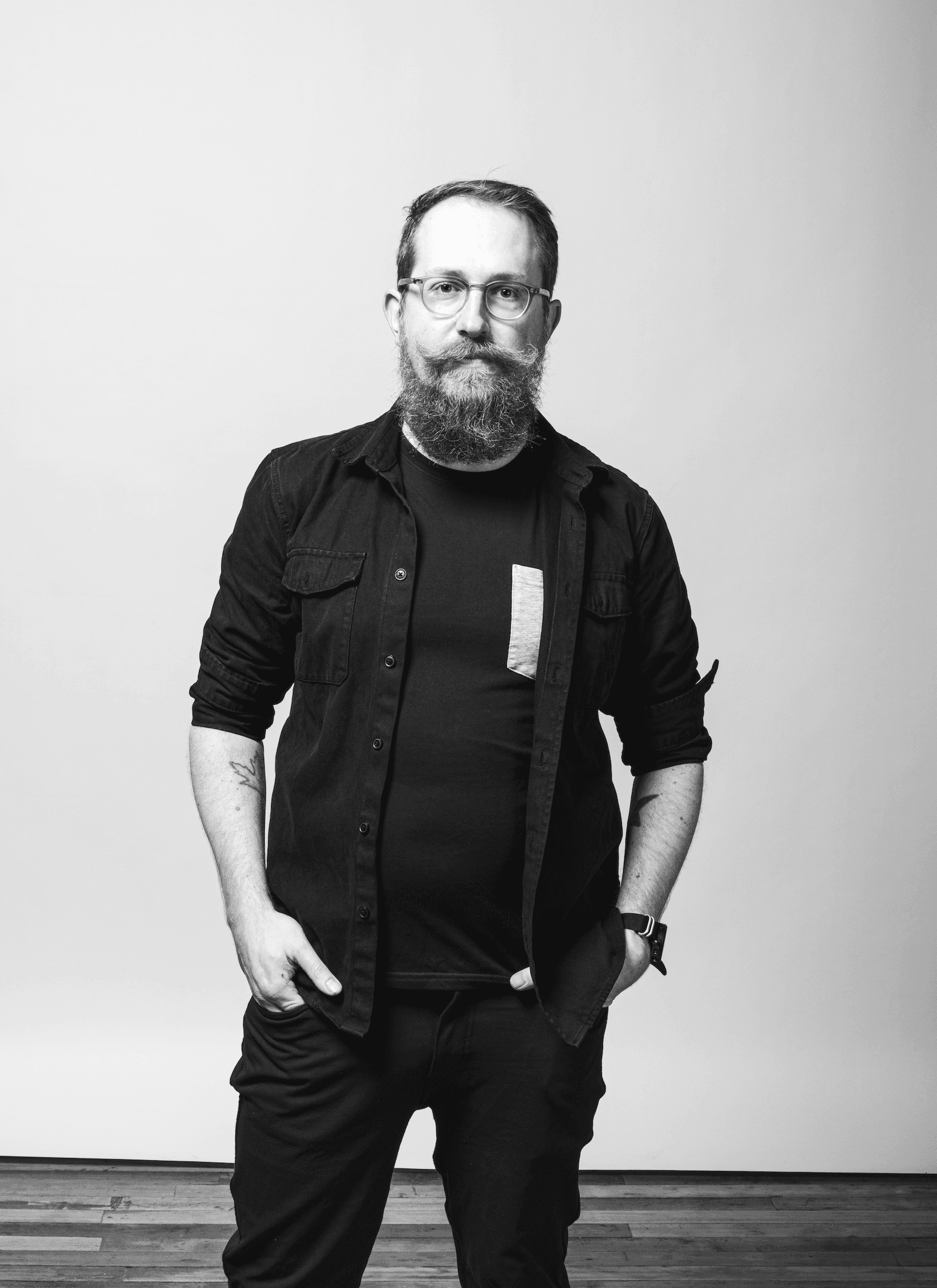 A black and white photo of a man standing in front of a white backdrop, wearing black clothes.